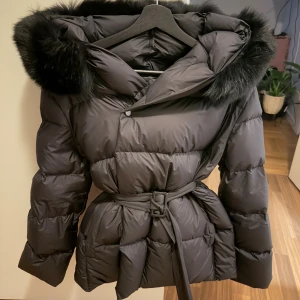 Svart dunjacka med päls från FUR Stockholm - Snygg svart dunjacka från FUR Stockholm med fluffig huva och pälsdetaljer. Jackan har bälte i midjan för en schysst siluett och är quiltad för extra värme. Perfekt för kalla dagar med sin långa ärm och stilrena look.