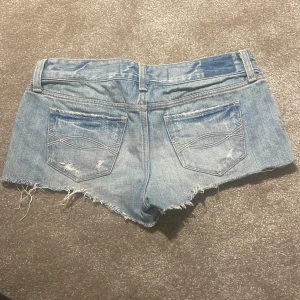 Blå lågmidjade jeansshorts Abercrombie & Fitch - Säljer ett par blå jeansshorts från Abercrombie & Fitch i klassisk denim med slitna detaljer och fransig kant. Shortsen har fem fickor, bälteshällor och låg midja. Perfekta för sommardagar och har en avslappnad, trendig look.