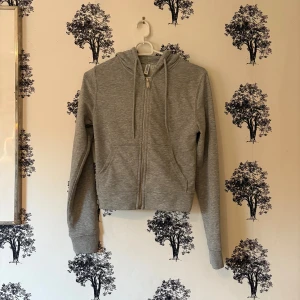Grå hoodie från H&M Divided  - Grå croppad hoodie från H&M Divided i storlek XS. Tröjan har huva med snörning, hel dragkedja framtill och två fickor. Mjuk bomullsmix och ribbade muddar vid ärmslut och nederkant. Perfekt för chill dagar eller när du vill ha en enkel look.
