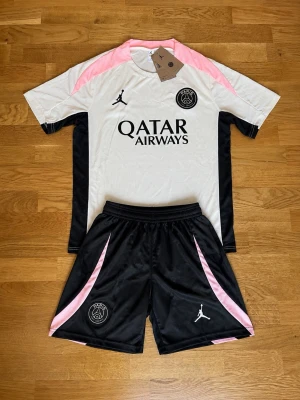PSG vit och rosa matchtröja - Snygg PSG matchtröja i vitt med svarta sidopaneler och rosa axelparti. Tröjan har korta ärmar, rund hals och tryck med Qatar Airways samt PSG-logga på bröstet. Tillverkad i lätt och ventilerande material, perfekt för fotboll eller träning.