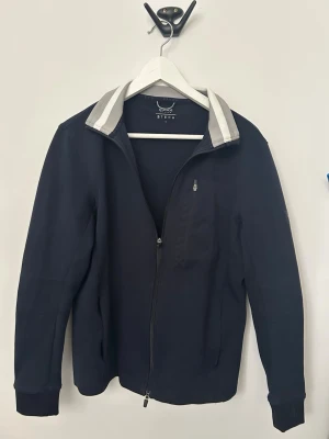 Svart jacka från Buoggi Milano BTECH - Stilren svart jacka från Buoggi Milano BTECH med dragkedja framtill och ståkrage med gråa detaljer. Jackan har en minimalistisk look och passar perfekt till vardagsoutfits. Tillverkad i ett mjukt material och har fickor på sidorna.