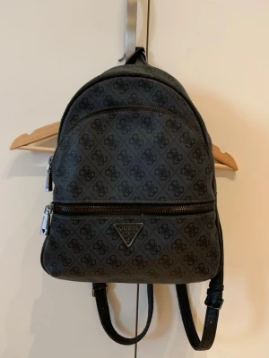 Mörkgrå ryggsäck från Guess - Snygg mörkgrå ryggsäck från Guess med klassiskt monogrammönster över hela väskan. Väskan har två stora fack med dragkedja, justerbara axelband och metalliska detaljer. Perfekt för dig som vill ha en trendig och praktisk accessoar.