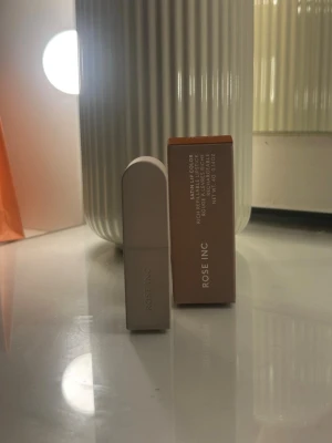 Rose Inc Satin Lip Color Besotted - Snyggt läppstift från Rose Inc i nyansen Besotted. Hylsan är stilren och matt beige med rundad topp. Läppstiftet är ett satin lipstick och kommer i en refillbar förpackning. Perfekt för dig som gillar en elegant look och vill ha ett modernt läppstift.