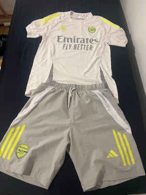Arsenal träningsset från Adidas - Snyggt Arsenal träningsset från Adidas med ljusgrå tröja och shorts. Tröjan har gula detaljer på axlarna, Arsenal-logga och 'Emirates Fly Better' tryck på bröstet. Shortsen är ljusgrå med gula Adidas-ränder och Arsenal-märke. Perfekt för fotbollsträning eller gym. Små skavanker finns har används lite under tiden annonsen varit publicerad. Fråga vid funderingar!