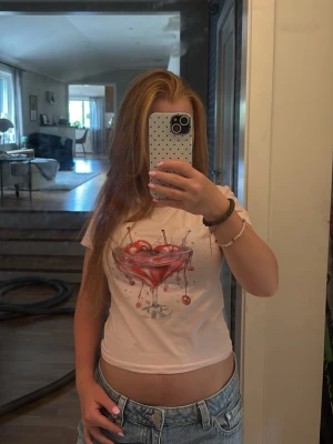 Ljusrosa croppad t-shirt med drinkmotiv - Säljer en ljusrosa croppad t-shirt med ett färgglatt tryck av en drink med körsbär på bröstet. T-shirten har korta ärmar och en avslappnad passform, perfekt för en trendig och chill look.