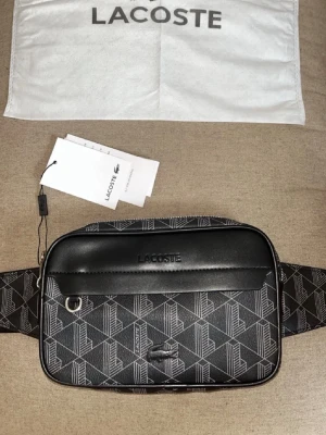 Svart magväska från Lacoste - Snygg svart magväska från Lacoste med geometriskt mönster och justerbart band. Väskan har en framficka med dragkedja, metallring för detaljer och präglad Lacoste-logga. Tillverkad i konstläder och textil, perfekt för att bära det viktigaste.