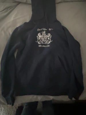 Mörkblå hoodie från Bvaldi - Mörkblå hoodie från Bvaldi med vit broderad logga och lejonemblem på bröstet. Klassisk huva med dragsko och känguruficka framtill. Perfekt för dig som gillar stilrena och sportiga plagg med en twist. Priset ej hugget i sten. Dm för frågor.