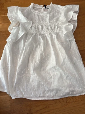 Vit kort klänning med volangärm - Söt vit kort klänning i bomull med volangärmar och diskret prickigt mönster. Klänningen har rund halsringning med liten volangkant och smockdetaljer över bröstet. Perfekt för sommarens alla tillfällen.