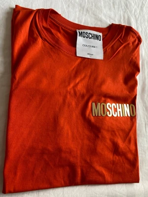 Moschino T-shirt - Snygg orange t-shirt från Moschino med rund halsringning och guldfärgad logga på bröstet. Tillverkad i mjuk bomull. Perfekt för dig som vill sticka ut med en exklusiv och trendig look.
