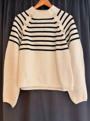 Vit stickad tröja med svarta ränder H&M - Säljer en vit stickad tröja från H&M med svarta horisontella ränder upptill och dekorativa guldknappar på axlarna. Tröjan har en hög hals och långa ärmar, perfekt för kyligare dagar. Materialet är tjockt och mjukt för extra komfort.