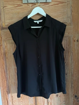 Svart ärmlös blus med krage - Stilren svart blus med klassisk krage och knäppning framtill. Blusen är ärmlös och har en rak, avslappnad passform som passar perfekt till både jeans och kjol. 