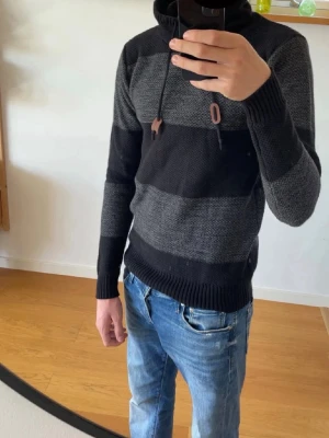 Stickad hoodie - Helt ny, Stickad hoodie med breda svarta och grå ränder, hög krage med dragsko och ribbstickade muddar. Perfekt för dig som gillar en avslappnad men ändå snygg stil. Materialet är mjukt och värmande, passar bra till jeans. Inköpt i Spanien 2026 från inside, säljer pga den är för liten. 