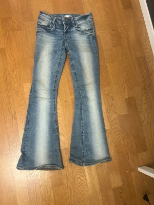 Blå bootcut jeans med slitningar - Säljer ett par blå bootcut jeans med snygga slitningar och ljus tvätt. Jeansen har klassiska fem fickor och utsvängda ben. Perfekta för dig som gillar retrostil och vill ha ett par jeans som sticker ut lite extra.