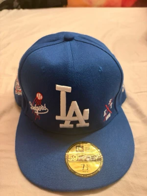 Blå LA keps - Snygg blå keps från New Era 59Fifty med LA Dodgers logga broderad framtill och detaljer på sidorna. Baksidan har texten 'Los Angeles' och MLB-logga. Klassisk flat peak och broderade ventilationshål. Perfekt för dig som gillar streetwear och sport. 