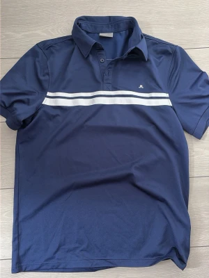 Marinblå piké från J.Lindeberg - Snygg marinblå pikétröja från J.Lindeberg med vit rand över bröstet och liten logga. Klassisk krage och korta ärmar. Perfekt till en avslappnad men stilren look.