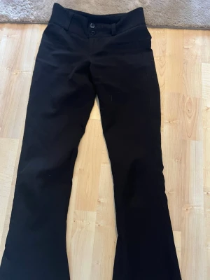 Svarta kostymbyxor med bootcut - Snygga svarta kostymbyxor med bootcut och låg midja. Byxorna har två knappar framtill, bälteshällor och fickor bak med lock och knapp. Perfekta för en clean och stilren look. Materialet ser ut att vara en mjuk och stretchig blandning.