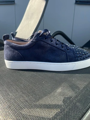 Mörkblå Loubs  - Säljer ett par mörkblå sneakers i mocka från Christian Louboutin. Skorna har coola svarta nitar på tån och vita sulor som ger en snygg kontrast. Klassisk låg modell med snörning och exklusiv känsla. Perfekta för dig som vill sticka ut med din stil.