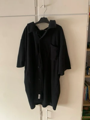 Oversized svart kappa med kort ärm - Säljer en oversized svart kappa med korta ärmar och knäppning framtill. Modellen har en bred krage och en bröstficka. Materialet ser ut att vara mjukt och lite tjockare, perfekt för lager på lager. Passar dig som gillar en avslappnad och trendig stil.