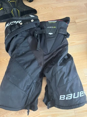 Bauer Hyperlite hockeybyxor svart - Säljer ett par svarta hockeybyxor från Bauer, modell Hyperlite. Byxorna har robust vaddering, justerbara spännen och slitstarkt material för extra skydd på isen. Perfekta för dig som vill ha lätta och skyddande byxor till hockeyträningen.