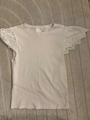 Ljusbeige topp med spetsärmar - Supersöt ljusbeige topp med ribbad struktur och korta, vida spetsärmar med broderade detaljer. Perfekt för dig som vill ha en feminin och trendig look. Rund halsringning och mjukt material.