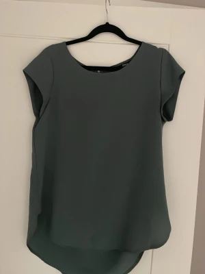 Mörkgrå/grön blus med dragkedja - Stilren mörkgrå/grön blus med rund halsringning och korta ärmar. Blusen har en snygg dragkedja i ryggen och lätt rundad nederkant. Passar även 38/40