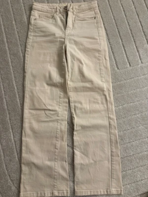 Beiga raka jeans med hög midja - Snygga beiga jeans med rak passform och hög midja. Klassisk femficksmodell med bälteshällor och knappgylf. Perfekta till vardagslooken och lätta att matcha med olika stilar. Materialet känns mjukt och bekvämt.