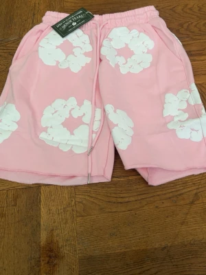 Rosa shorts med vita blommor - Säljer ett par ljusrosa shorts med vita blommönster och resårmidja med snörning. Shortsen har sidofickor och är tillverkade i mjukt bomullstyg. Perfekta för varma dagar och ger en avslappnad vibe.