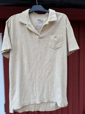Beige frotté t-shirt från OAS - Säljer en beige t-shirt från OAS i mjuk frotté. T-shirten har korta ärmar, öppen krage och en bröstficka med diskret broderi. Perfekt för en avslappnad och chill stil. Passar dig som gillar unika material och enkel design.
