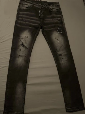 Svarta skinny jeans med slitningar - Säljer ett par svarta skinny jeans med slitningar och blekta detaljer på benen. Jeansen har fem fickor, en metallring framtill och en patch med texten 'Caten Treated' bak. Snygg streetstil och coola detaljer som passar perfekt till en trendig look.