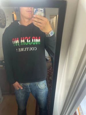 Svart Moschino hoodie  - Jag är 186cm. Säljer en svart hoodie från Moschino med färgglatt tryck i rött, vitt och grönt där det står 'MOSCHINO COUTURE!'. Hoodien har huva med dragsko och ribbade muddar. Perfekt för dig som vill sticka ut med en ikonisk streetwear-look.