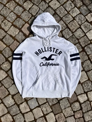 Vit Hollister hoodie med tryck - Säljer en vit hoodie från Hollister med marinblå tryck på bröstet där det står 'Hollister California' och en stor logga. Hoodien har huva med dragsko, känguruficka och två marinblå ränder på varje ärm. Perfekt för en avslappnad och sportig stil.