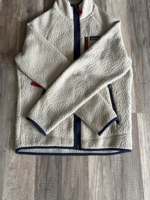 Beige fleecejacka från Patagonia - Mysig beige fleecejacka från Patagonia med marinblå detaljer och dragkedjor. Jackan har hög krage, tre fickor med dragkedja och röda zip-pullers. Perfekt för lager-på-lager och outdoor-vibe.