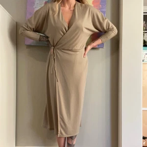 Beige omlottklänning med knytband - Stilren beige omlottklänning med v-ringning och långa ärmar. Klänningen har ett knytband i midjan som ger en snygg siluett och är tillverkad i ett mjukt, stretchigt material. Perfekt för dig som gillar enkel men elegant stil.