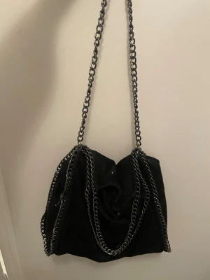 Svart axelväska med kedjedetaljer - Cool svart axelväska i mjukt material med kedjor som axelrem och runt kanterna. Väskans tyg är lite lätt glittrigt/skimrigt. Perfekt för dig som gillar coola accessoarer och vill ha plats för allt du behöver i väskan.