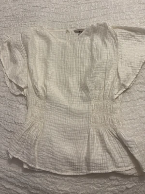Vit blus från H&M - Säljer en vit blus från H&M i storlek S. Blusen har en strukturerad, krinklad bomullskänsla, korta ärmar och smockdetaljer i midjan som ger en snygg siluett. Perfekt till jeans eller kjol för en fräsch look.