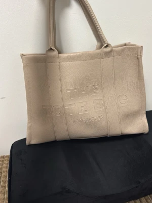 Beige väska - Säljer en beige  i skinn med präglad logotyp på framsidan och en patch på baksidan. Väskan har två rejäla handtag och en rymlig, boxig form som gör den perfekt för att bära allt du behöver. Stilren och trendig design. 