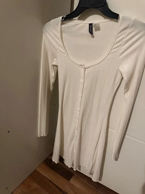 Vit ribbad kortklänning med knappar - Säljer en vit ribbad kortklänning med lång ärm och knappar framtill. Klänningen har en rundad urringning och är tillverkad i ett stretchigt material som sitter snyggt på kroppen. Perfekt för dig som gillar enkel men stilren design.