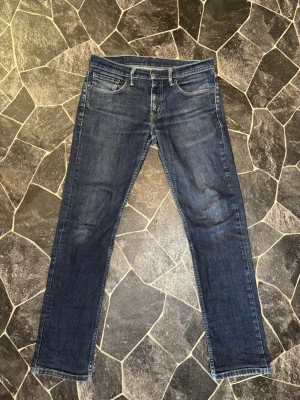 Levi's 511 blå jeans slim fit - Säljer ett par klassiska blå Levi's 511 jeans i slim fit-modell. Jeansen har fem fickor, dragkedjegylf och snygga slitningar framtill. Tillverkade i bomull med en liten mängd stretch för extra komfort. Perfekta till en avslappnad stil.