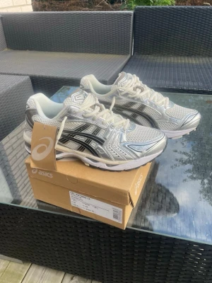 ASICS GEL-Kayano 14 silver/vita sneakers - Snygga ASICS GEL-Kayano 14 sneakers i silver och vitt med svarta detaljer. Skorna har en sportig mesh- och syntetöverdel, klassisk snörning och chunky sula. Perfekta för dig som gillar retrovibbar och streetstyle.