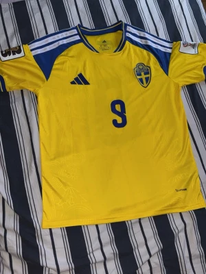 Svensk landslagströja nummer 9 - Gul och blå fotbollströja från Adidas med Sveriges landslagsemblem på bröstet och nummer 9 både fram och bak. Namnet 'ISAK' står på ryggen. Tröjan har korta ärmar, rund hals och Climacool-material som andas. Perfekt för match eller träning.
