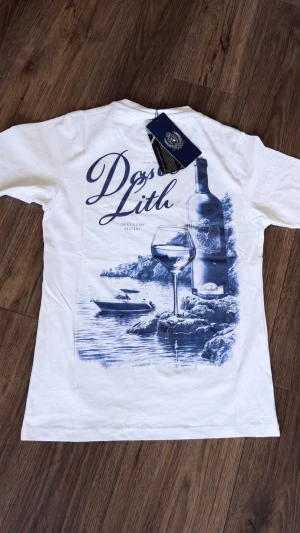 Vit t-shirt med vintagemotiv - Vit t-shirt med stort blått tryck på ryggen som föreställer en vinflaska, ett vinglas och en båt vid vattnet. Klassisk passform och rund hals. Perfekt för dig som gillar unika och stilrena motiv.