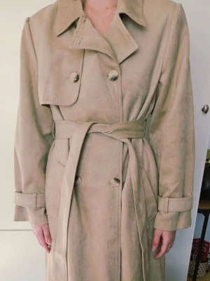 NA-KD beige trenchcoat, storlek EU 42 - Helt aldrig använd. Stilren beige trenchcoat med dubbelk. Tillverkad i ett mjukt material som ger en lyxig känsla. En lång trenchcoat med dubbelknäppning och ett matchande knytbälte i midjan. Kappan har klassiska detaljer som slag, axelklaffar och manschetter med spännen. En slits baktill ger extra rörelsefrihet. Köparen står för frakt. Ej retur.