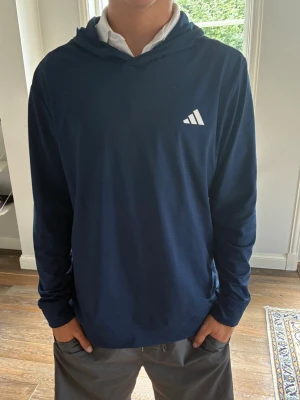 Mörkblå hoodie från Adidas - Snygg mörkblå hoodie från Adidas med vit logga på bröstet. Hoodien har huva och långa ärmar, tillverkad i ett mjukt och lätt material som passar perfekt till en avslappnad stil. Enkel och stilren design utan fickor eller dragkedja. Perfekt att ha på golfbanan eller på gymmet 