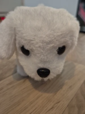 Vit mjuk leksakshund - Söt vit mjuk leksakshund med svarta ögon och nos. Pälsen är fluffig och mjuk, perfekt som gosedjur eller dekoration. Hunden har hängande öron och en rund form som gör den extra gullig man kan trycka på en knapp på undersidan av hunden för att starta den så den går köpt på marknad