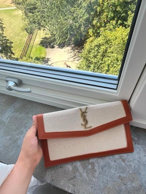 Beige kuvertväska med bruna detaljer - Stilren kuvertväsk i beige canvas med bruna läderdetaljer och guldfärgad- på framsidan. Väskan har en klassisk rektangulär form och stängs med lock. Perfekt accessoar för en trendig look.