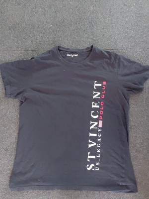Svart t-shirt US Legacy Polo Club - Snygg svart t-shirt med vertikal text 'ST VINCENT US LEGACY POLO CLUB' i vitt och rött framtill. Klassisk rund hals och korta ärmar. Perfekt till jeans eller shorts för en avslappnad look.Står XL men passar L och M
