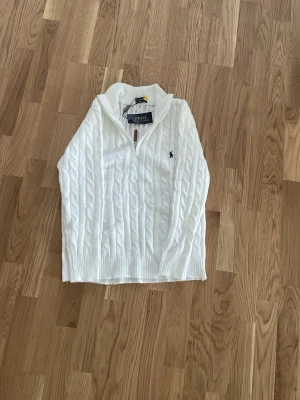 Vit kabelstickad tröja Polo Ralph Lauren - Kabelstickad vit tröja från Polo Ralph Lauren med halv dragkedja och hög krage. Tröjan har det klassiska broderade logotypen på bröstet och ribbade muddar vid ärmslut och nederkant. Perfekt för en stilren och avslappnad look