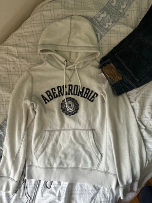 Grå hoodie från Abercrombie - Snygg grå hoodie från Abercrombie & Fitch med stor logga och emblem på bröstet. Klassisk känguruficka framtill, dragsko i huvan och mjukt material. Perfekt för en avslappnad och trendig look.