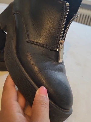 Svarta stövletter med dragkedja - Säljer ett par svarta stövletter i skinn med chunky sula och grov dragkedja framtill. Skorna har en modern, kantig design och är perfekta för dig som gillar en edgy stil. Passar till både jeans och kjol för en cool look. Från Zara. Storlek 37!