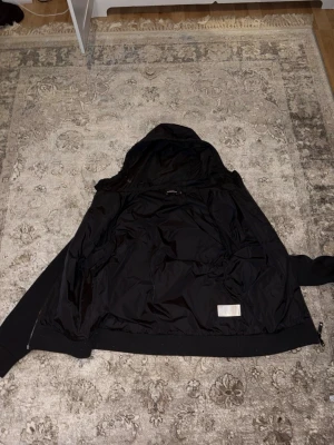 Svart J.Lindeberg cardigan jacka med huva - 2 små hål i luvan syns ej vid användning.! Svart cardigan jacka med huva och dragkedja. Kroppen är vadderad i glansigt material medan ärmar och muddar är stickade. Jackan har en bröstficka med knapp och två sidofickor. Perfekt för en chill och trendig look.